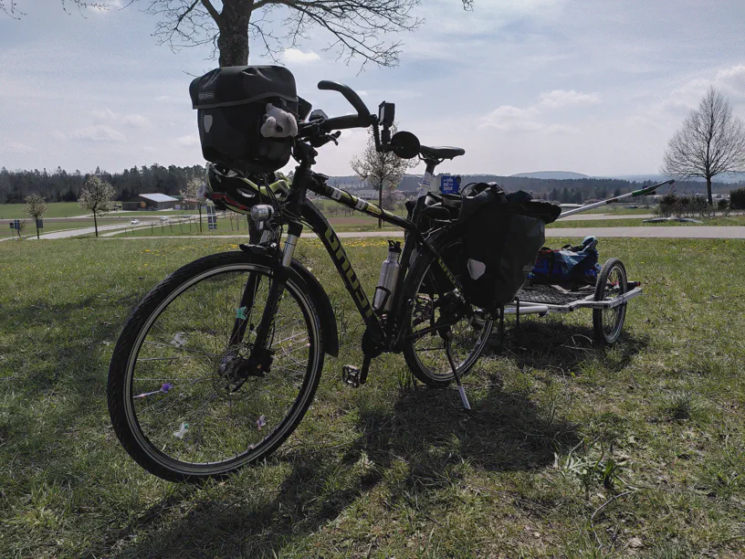 Auf einer Wiese steht ein Fahrrad mit einem Anhänger. In der Lenkertasche sitzt der kleine Elefant Kagube und hinten sieht man den Fahrradanhänger Ploggenom.