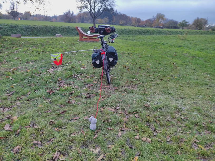 Kleiner Elefant sitzt auf dem grünen Schlitten auf der Wiese und die Schnur führt zum dahinter stehenden Fahrrad vom Radfahrer. Szene von weiter oben als beim vorherigen Bild.