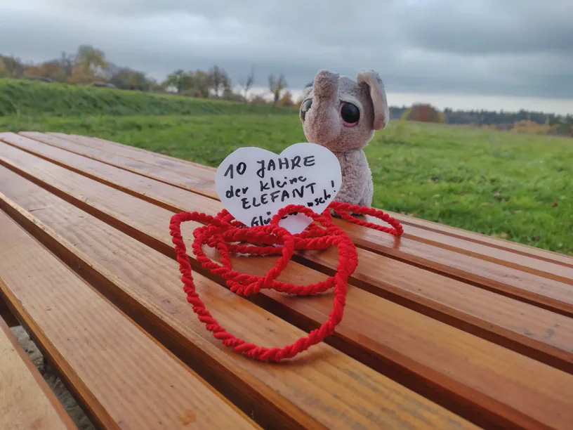 Kleiner Elefant sitzt neben einem beschriebenen Herzchen, auf dem steht: '10 Jahre der kleine Elefant – Glückwunsch'.
