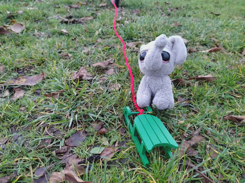 Kleiner Elefant sitzt neben dem grünen Schlitten auf der Wiese und da ist eine Schnur dran und die führt aus dem Bild raus.