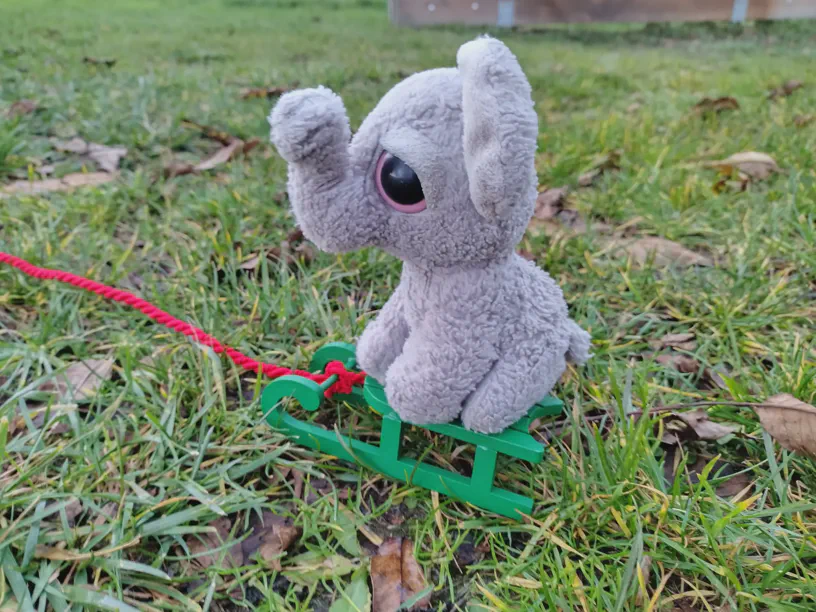 Kleiner Elefant sitzt auf dem grünen Schlitten auf der Wiese und da ist eine Schnur dran und die führt aus dem Bild raus.