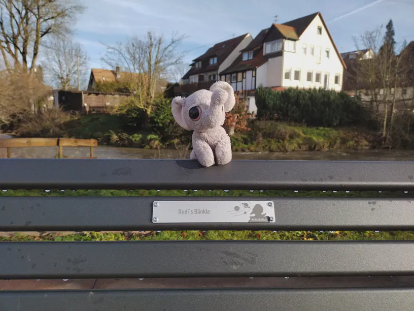 Kagube sitzt auf der Rückenlehne einer Sitzbank und ein Schild daran zeigt 'Rudi‘s Bänkle'. Unten rechts steht Kepplerstadt, dazu ein paar Planeten und ein Kopf mit Bart.